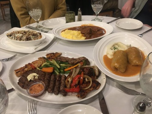 A table set with various dishes, including grilled meats, mashed potatoes, and stuffed peppers, surrounded by glasses.