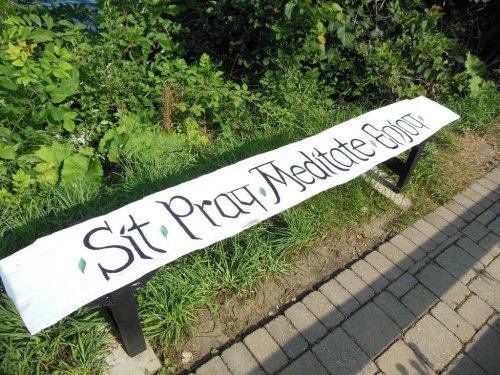 A wooden bench with the words "Sit Pray Meditate" painted on it, surrounded by greenery.