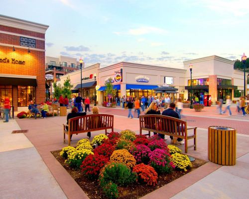 A vibrant outdoor shopping area with benches, colorful flowers, and people walking around shops at sunset.