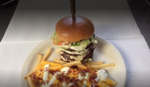 A large burger with multiple patties, lettuce, and tomato, served with cheesy bacon fries on a plate.