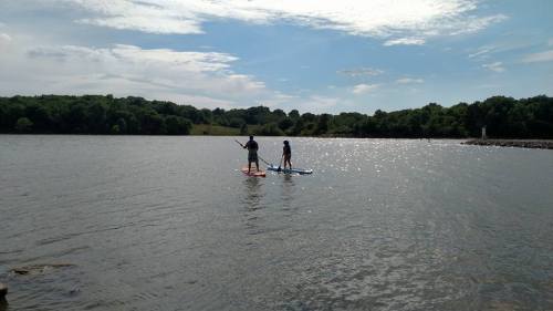Two people paddleboarding on a calm lake, surrounded by greenery and a partly cloudy sky.