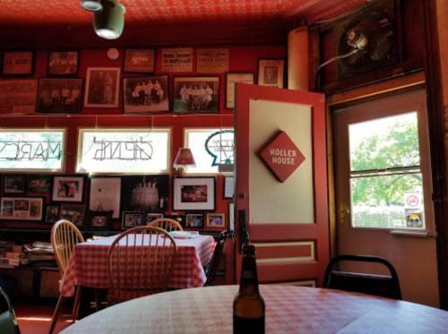 Cozy restaurant interior with red walls, checkered tablecloths, and vintage photos. A door labeled "Holler House" is visible.