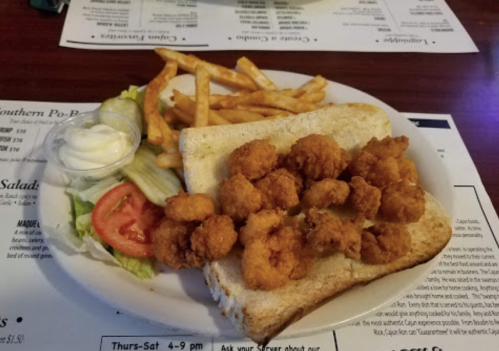 A plate featuring fried shrimp on bread, served with lettuce, tomato, a side of fries, and a small cup of sauce.