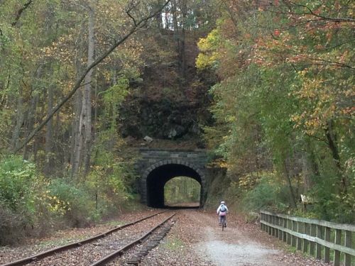 A cyclist rides along a trail leading to a stone tunnel, surrounded by autumn foliage and trees.