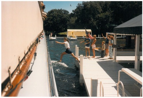 A group of people on a dock watch as a girl jumps from a boat into the water on a sunny day.