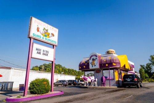 A colorful fast-food restaurant named "Purple Cow" with a sign announcing "Mac Bites Are Back." Cars are parked outside.