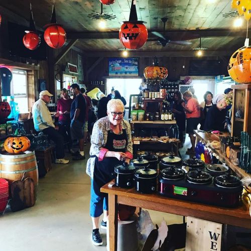 A busy store decorated for Halloween, with a smiling woman in an apron serving food among customers and festive decor.