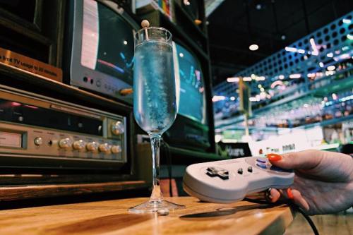 A hand holds a game controller next to a glass of blue drink, with vintage TVs in the background.