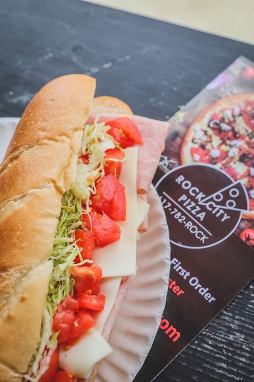 A sub sandwich filled with lettuce, tomatoes, and cheese, next to a menu for Rock City Pizza.