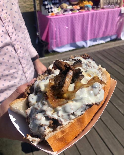 A hand holding a loaded sandwich with beef, mushrooms, onions, and creamy sauce, set against a festive background.