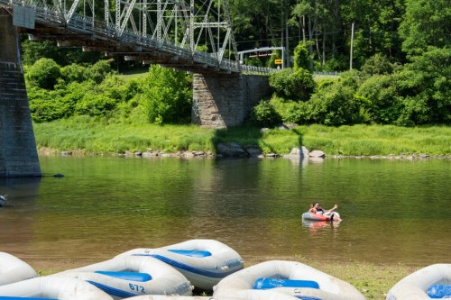 Two people relax in a small inflatable boat on a calm river, with a bridge and green trees in the background.