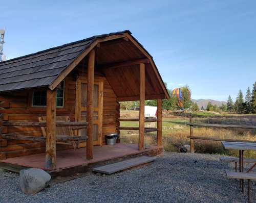 A rustic log cabin with a porch, surrounded by nature and a colorful hot air balloon in the background.