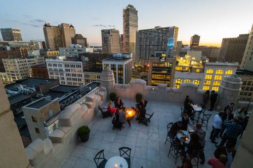 A rooftop bar scene at sunset, with people socializing around fire pits and city skyline in the background.