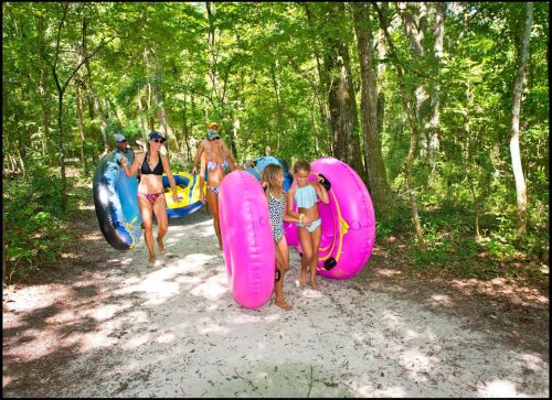 A group of people, including children, carry colorful inner tubes along a sandy path in a lush, green forest.