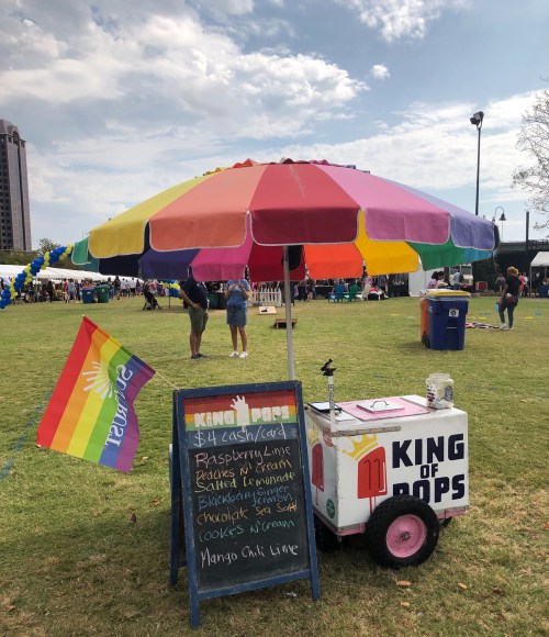 A colorful ice pop cart with a rainbow umbrella, featuring a menu board and a pride flag in a park setting.