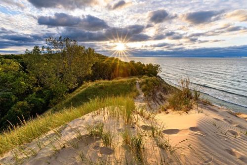 Sunset over a sandy dune with grass, overlooking a calm lake and cloudy sky.