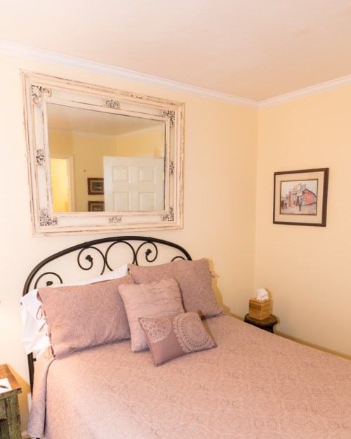 Cozy bedroom with a bed, decorative pillows, a vintage mirror, and a framed picture on the wall. Soft yellow walls.