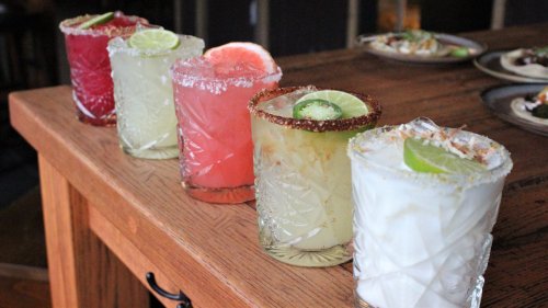 Five colorful cocktails in decorative glasses, garnished with lime and other fruits, arranged on a wooden table.