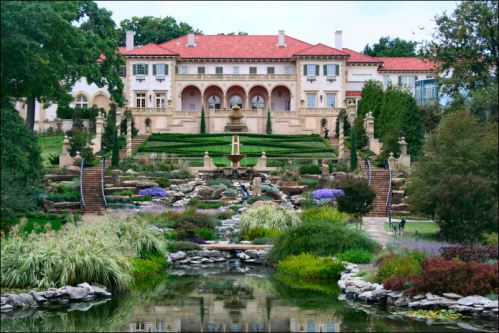 A grand mansion with a red-tiled roof, surrounded by lush gardens, fountains, and a serene pond in the foreground.