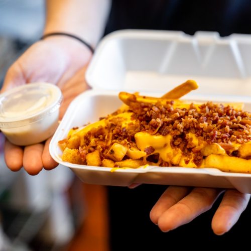A person holds a container of loaded fries topped with cheese and bacon, alongside a small cup of dipping sauce.