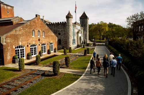 A group of people walks along a path near a castle-like building and landscaped gardens on a sunny day.