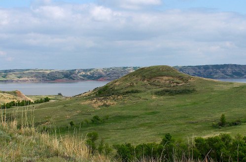 A grassy hill overlooks a calm lake, with rolling hills and cloudy skies in the background.