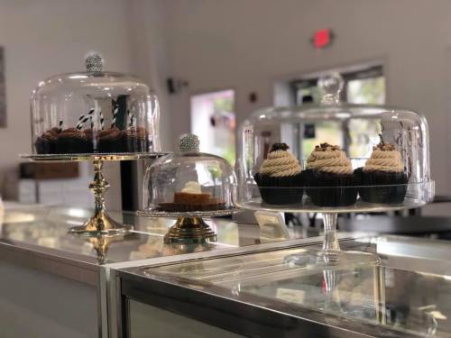 Three glass domes display an assortment of cupcakes and desserts on a counter in a bright bakery.