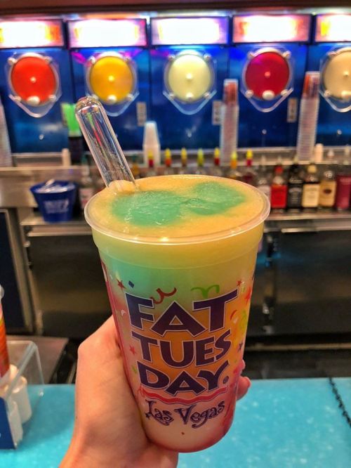 A colorful frozen drink in a cup labeled "Fat Tuesday" held in front of a slushy machine with various flavors.