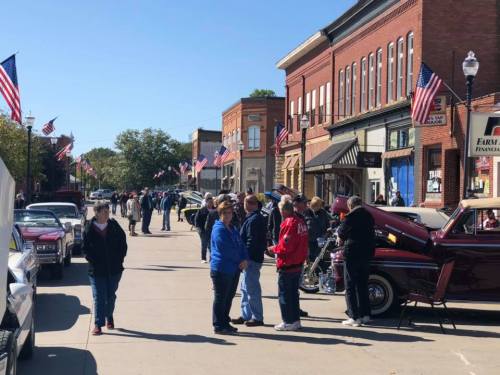 A lively street scene with people gathered near classic cars, flags lining the buildings, and a clear blue sky.