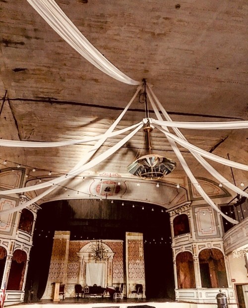 Interior of a vintage theater with draped fabric, a chandelier, and a stage set for an event.