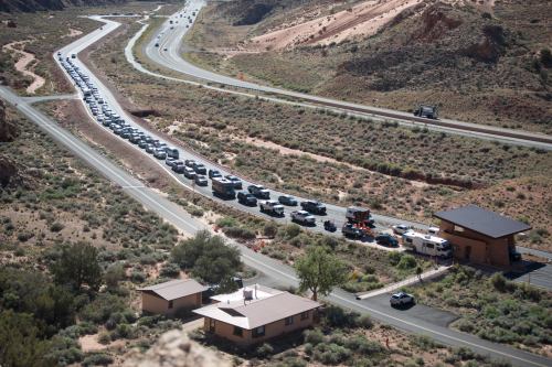 A long line of cars on a winding road in a desert landscape, with buildings and greenery nearby.