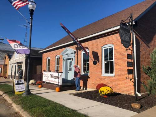 A brick building with a "Welcome Center" sign, colorful flowers, and a person standing outside on a sunny day.