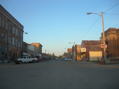 A quiet street scene with old buildings, parked cars, and clear blue sky in a small town.