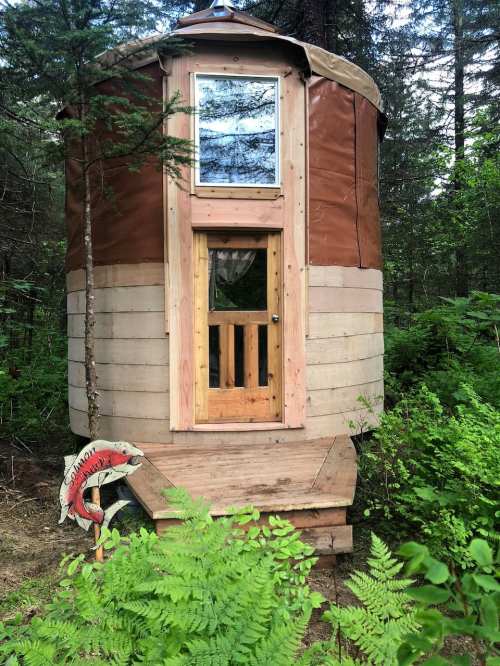 A small, round cabin with a wooden door and window, surrounded by trees and greenery. A life preserver is nearby.
