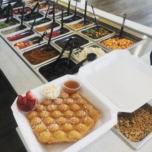 A waffle topped with strawberries and white chocolate, alongside a variety of toppings in containers.