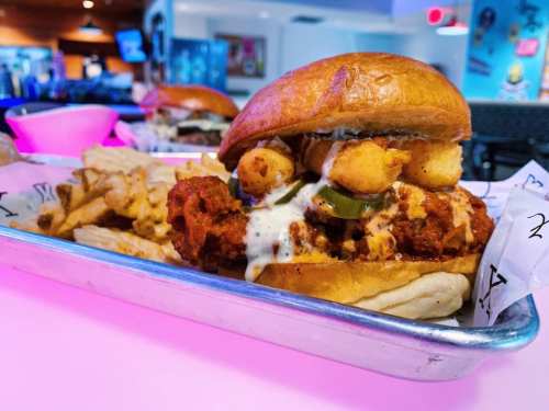 A close-up of a crispy chicken sandwich topped with pickles and sauce, served with crinkle-cut fries on a tray.