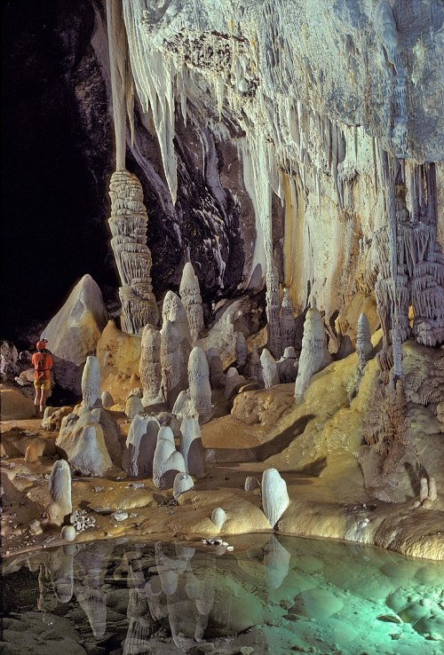 A person stands in a cave surrounded by stalagmites and stalactites, with a reflective pool of water nearby.
