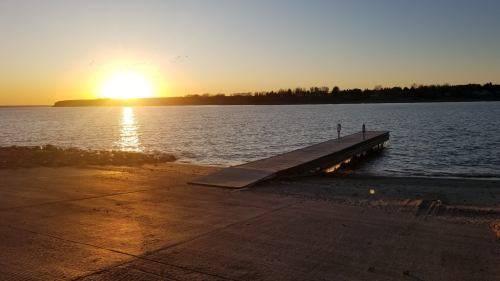 Sunset over a calm lake, with a wooden dock extending into the water and silhouettes of trees in the background.