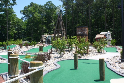A scenic mini-golf course featuring green turf, a windmill, waterfall, and rustic buildings surrounded by trees.