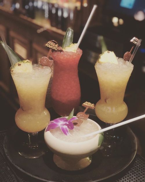Four colorful cocktails with garnishes, served in elegant glasses on a tray, set against a dimly lit bar background.