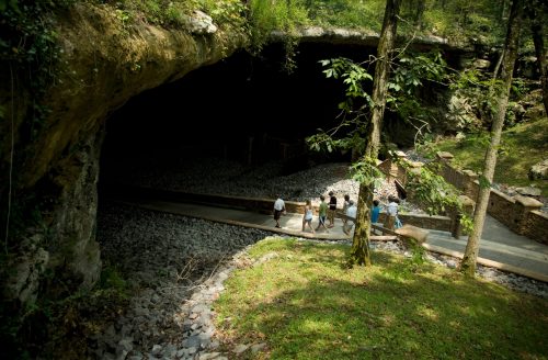 Visitors walk along a path leading to a large cave entrance surrounded by trees and rocky terrain.