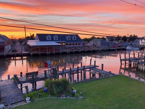 A serene waterfront scene at sunset, with colorful skies reflecting on calm water and boats docked along the pier.
