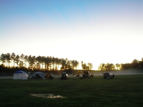 A serene campsite at dawn, featuring tents and a vehicle, with mist rising over a grassy field and trees in the background.