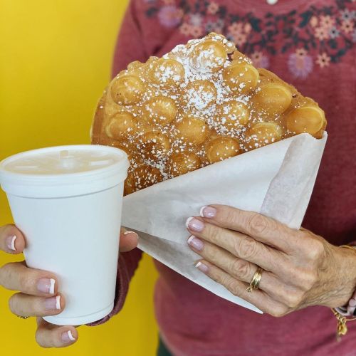 A person holds a large bubble waffle topped with powdered sugar and a white cup, set against a bright yellow background.
