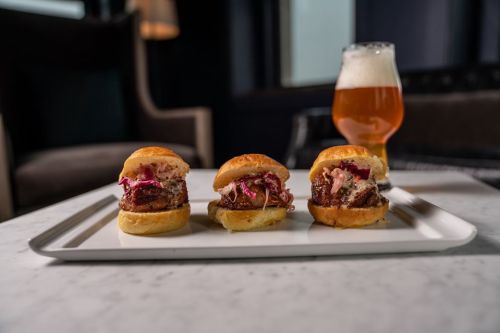 Three sliders on a white plate, accompanied by a glass of beer, set against a dark, elegant background.