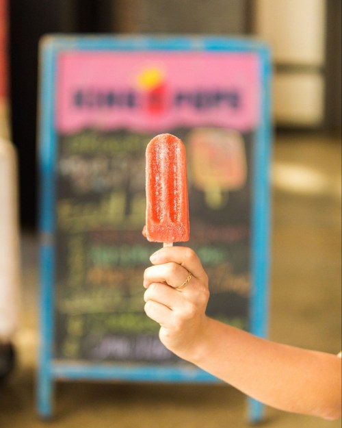 A hand holding a red popsicle in front of a colorful chalkboard sign.