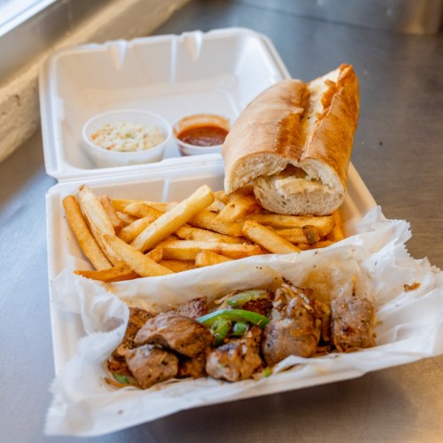 A takeout container with grilled meat skewers, fries, a sandwich, and two dipping sauces.