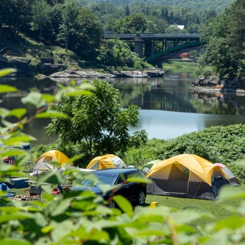 A scenic campsite by a river, featuring yellow tents, a car, and lush greenery, with a bridge in the background.