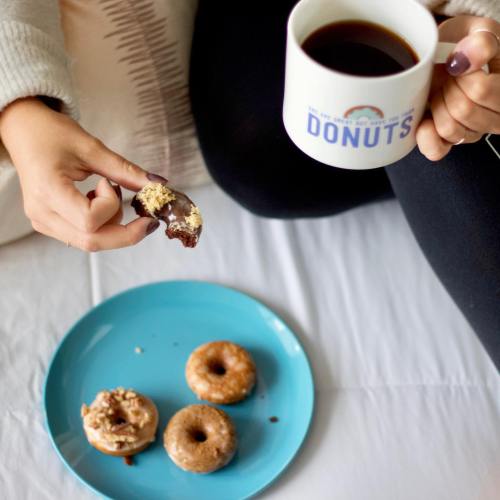 A person holds a coffee mug and a half-eaten donut, with a plate of assorted donuts nearby.