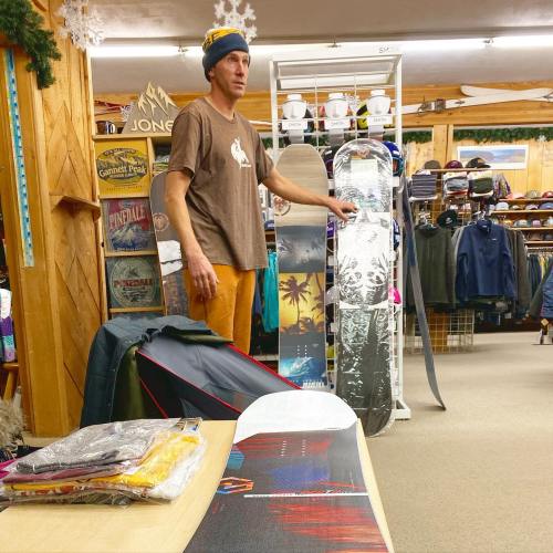A man stands in a ski shop, surrounded by snowboards and winter apparel, with a display of gear in the background.
